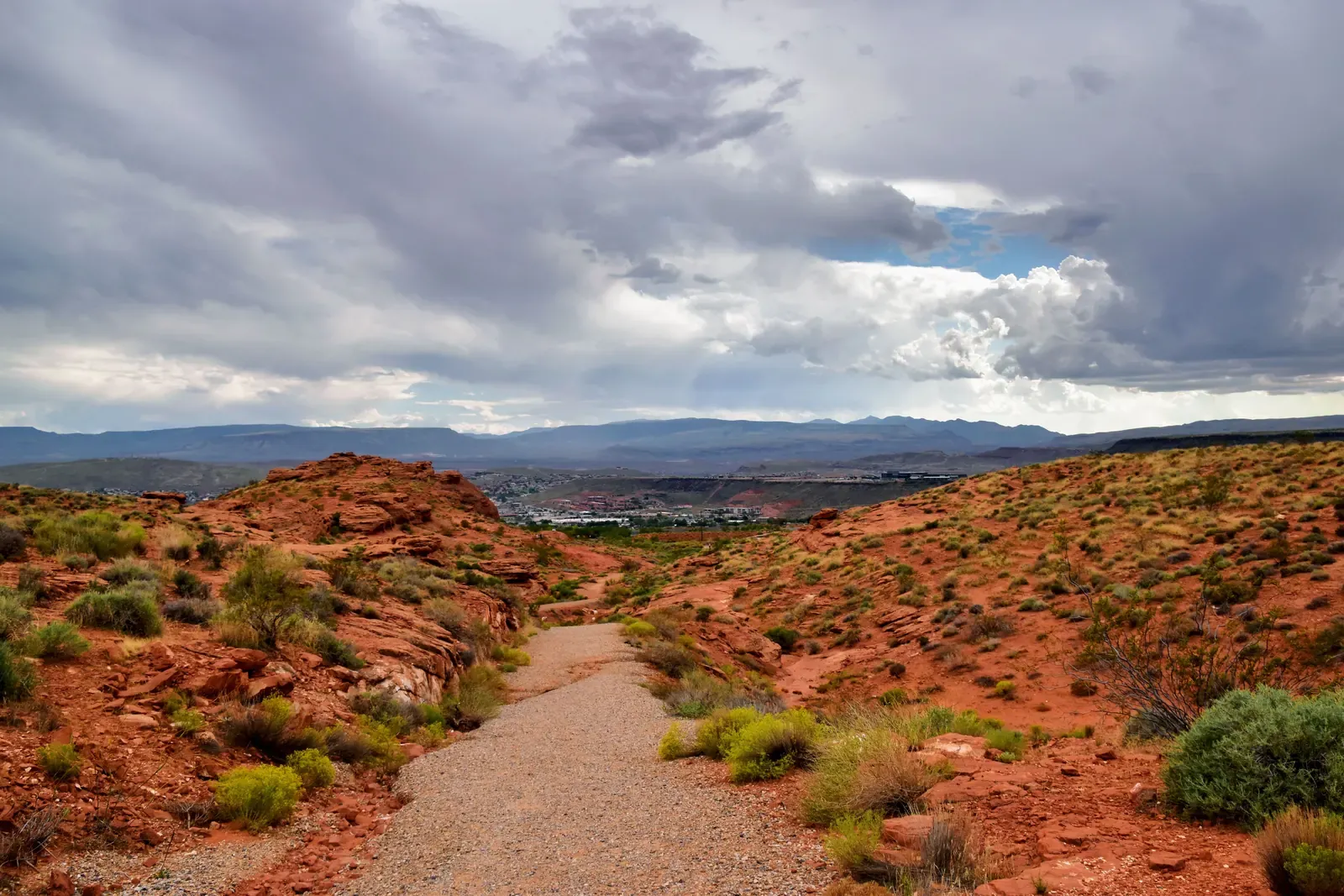 Gravel trail through red rock and desert shrubs above the St. George, Utah valley under a cloudy sky